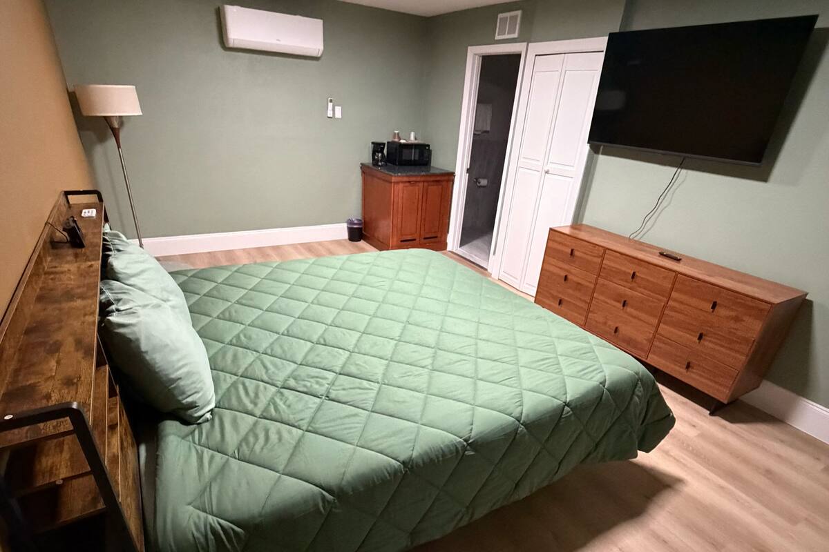 A bedroom with a bed, a wooden dresser, a television mounted on the wall, and a door leading to another room.