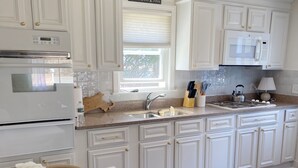 A kitchen with white cabinetry, a built-in oven, a microwave, and a window with blinds. - Cape Cod Retreat - Sleeps 6 - A/C & Outdoor Shower (Falmouth)