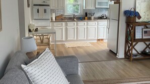 A modern kitchen with white cabinets, a central island, and a dining area with a gray sofa and striped pillows. - Cape Cod Retreat - Sleeps 6 - A/C & Outdoor Shower (Falmouth)