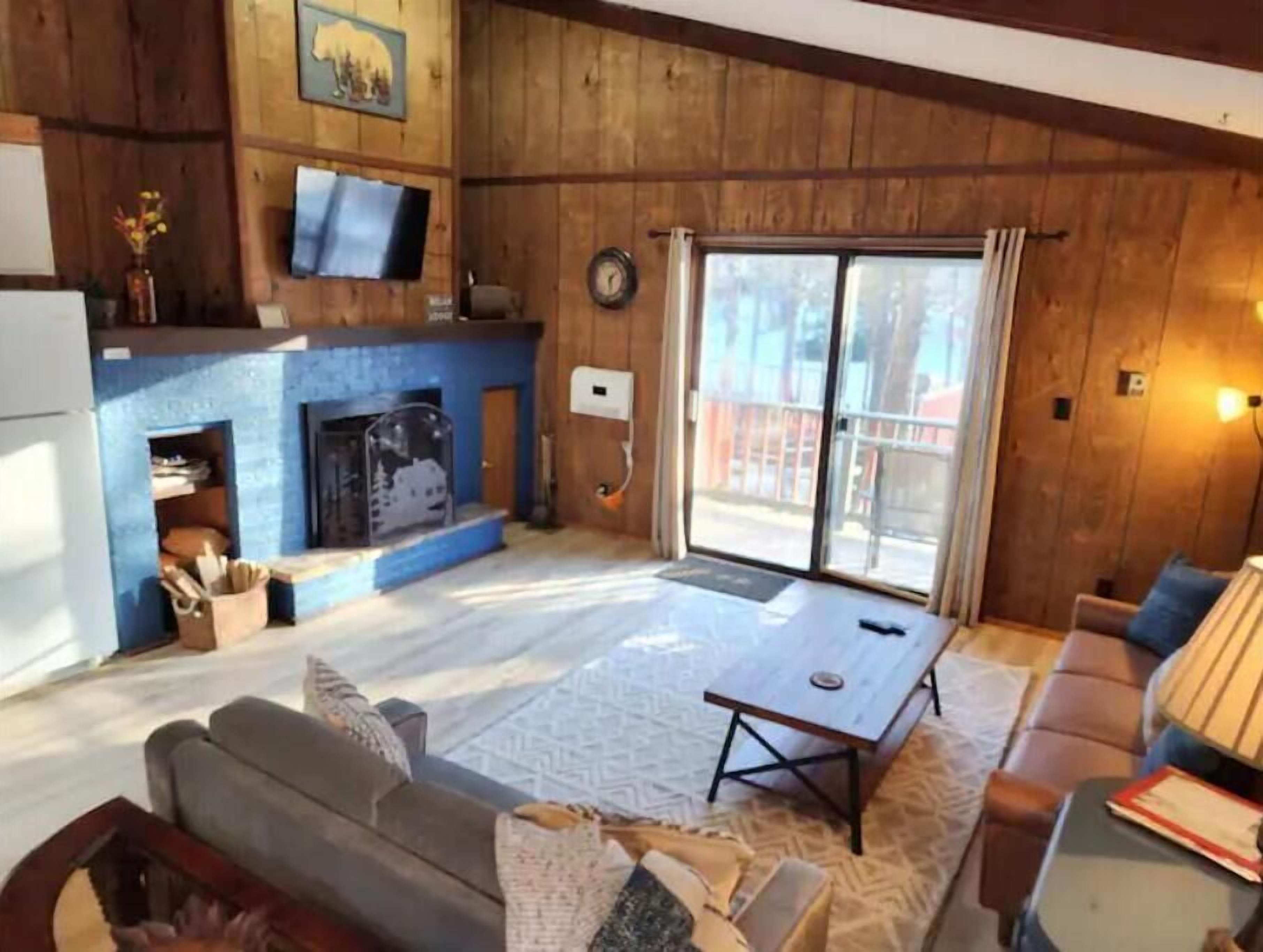 A cozy living room with a fireplace, a television mounted on the wall, and a large sliding glass door leading to an outdoor area.