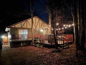 A cabin with a deck, surrounded by trees and lit by string lights.