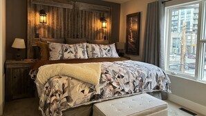 Ein Schlafzimmer mit einem großen Bett, einem Holz-Kopfteil, zwei Wandlampen, einem Fenster mit Blick auf Gebäude und einem Nachttisch mit einer Lampe.