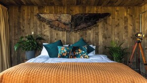 Ein Schlafzimmer mit Holzboden, einem großen Bett mit orangefarbener, strukturierter Decke und zwei grünen Kissen. In der Ecke steht eine Kamera auf einem Stativ.