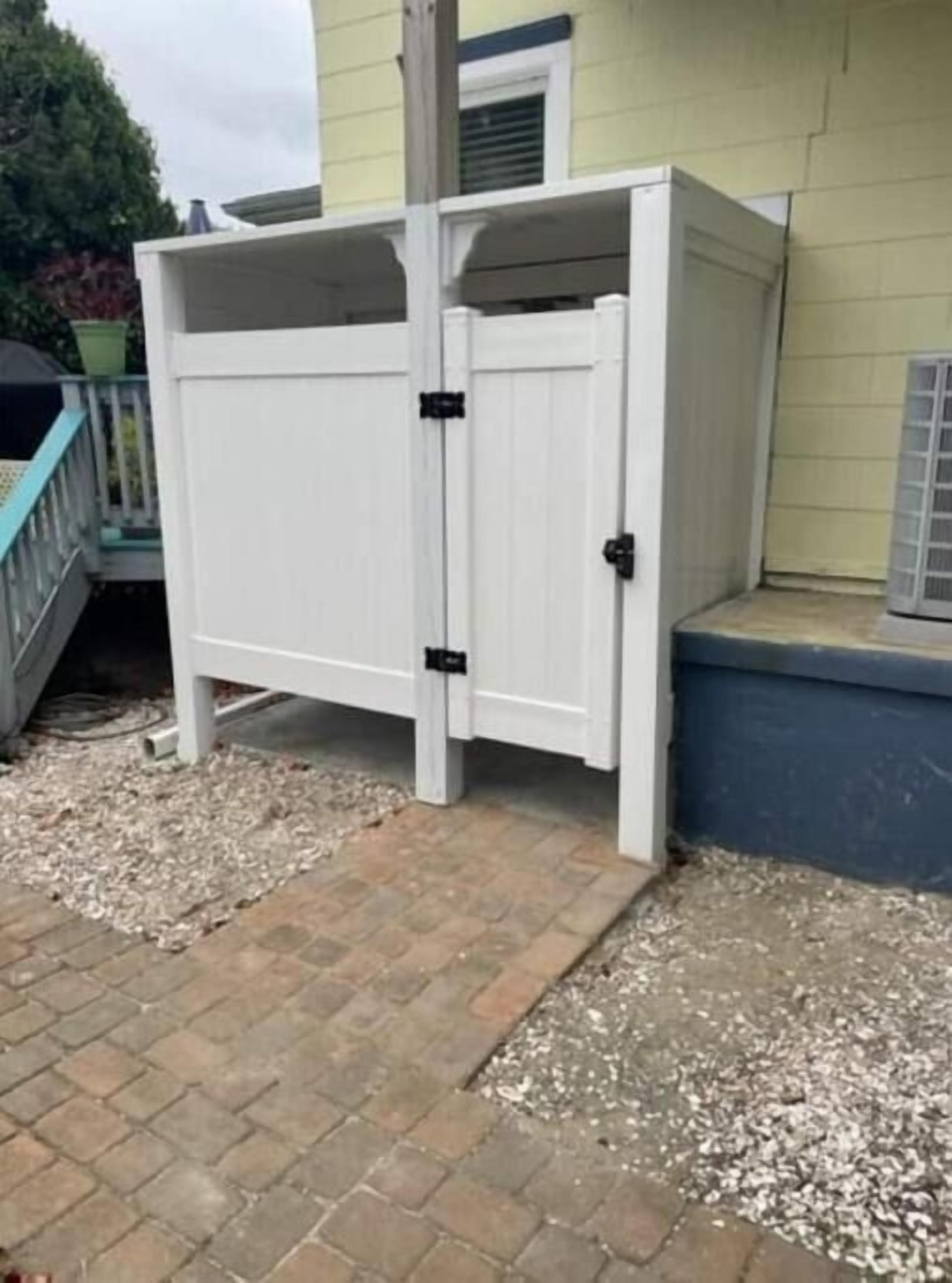 A white outdoor storage unit with a hinged door, positioned on a brick patio adjacent to a house with a blue trim and a white door.