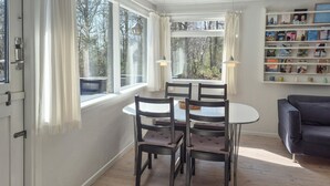 Ein Essbereich mit einem runden Tisch und vier Stühlen, ein Bücherregal mit Büchern und Blick auf Bäume durch große Fenster.
