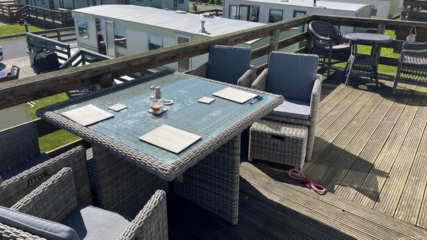 A deck with wicker furniture and a table, overlooking a coastal area with mobile homes and a view of the sea. - 3 Bedroom Lodge, Close to Beach, Stunning Views, includes hot tub (Drummore)