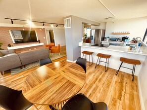Un soggiorno moderno con un tavolo da pranzo in legno, un divano grigio e poltrone arancioni. Si intravede anche un angolo cottura con frigorifero e sgabelli da bar.