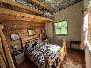 A rustic bedroom with a wooden bed, a nightstand, a lamp, a window with a view of greenery, and a ceiling with a wooden beam.
