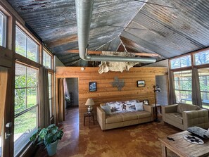 A rustic interior with wooden walls, a leather sofa, a coffee table, and a large window offering a view of greenery.