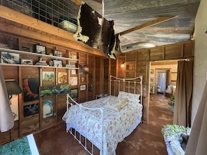 A rustic wooden room with a bed, shelves with various items, and a cowhide on the ceiling.