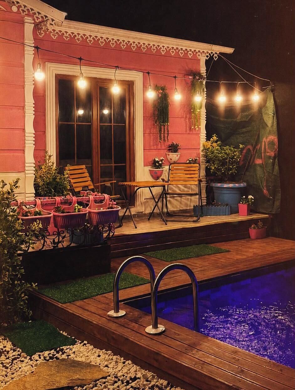 A poolside area with a wooden deck, a pool with a blue tint, outdoor seating, and string lights.