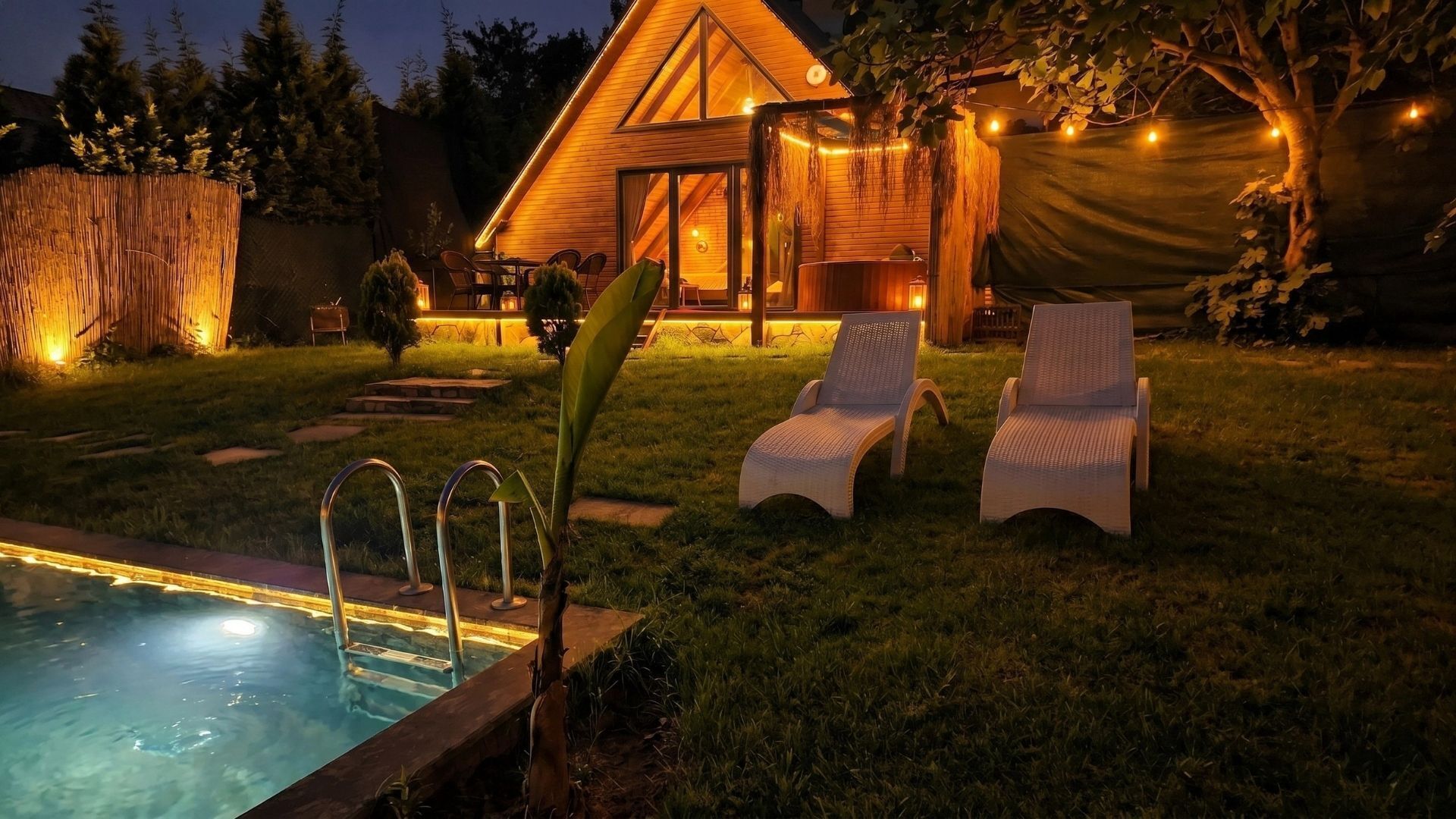 Un jardin avec une piscine, deux chaises longues et un abri en bois illuminé de guirlandes lumineuses.