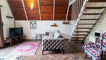 Habitación estilo loft con techo de madera, un sofá, una silla, un televisor, una pequeña mesa y una escalera.