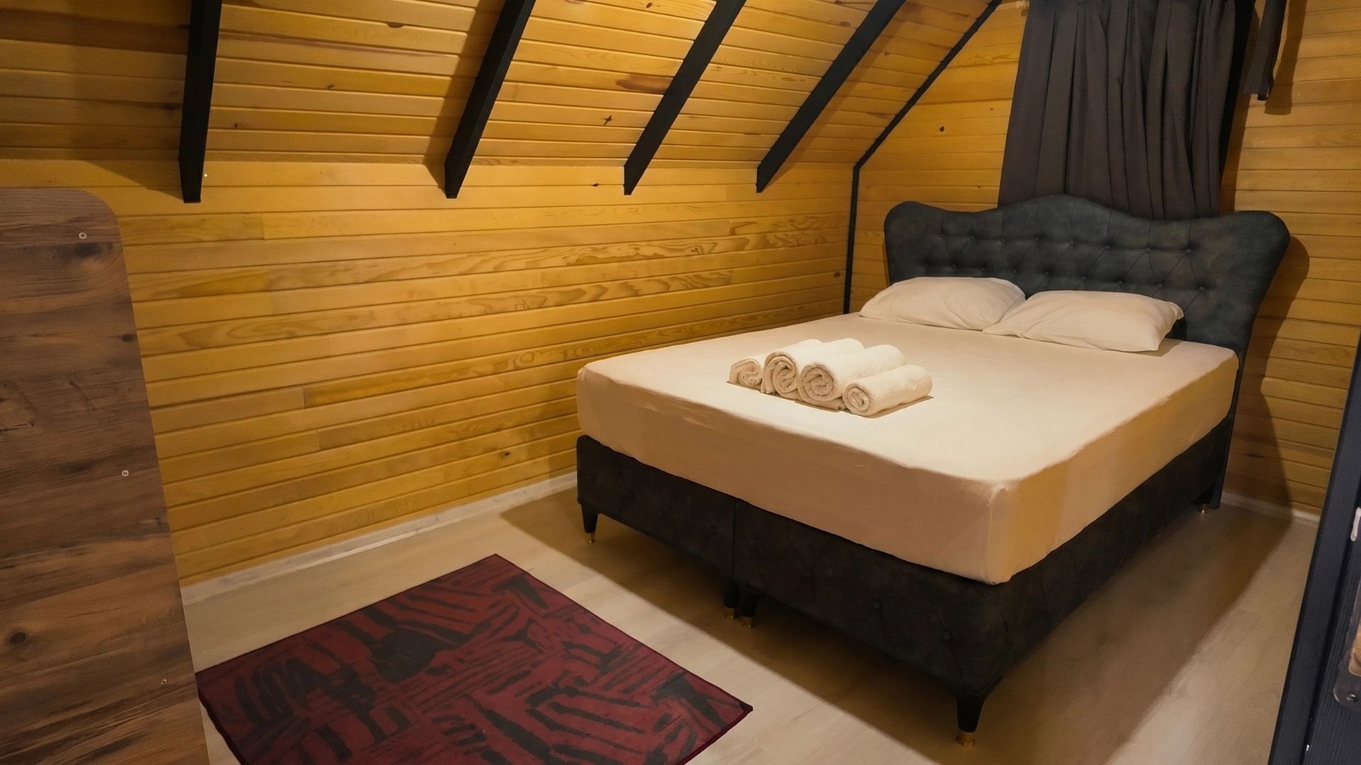 A bedroom with a wooden sloped ceiling, a bed with a dark headboard, and a red patterned rug.