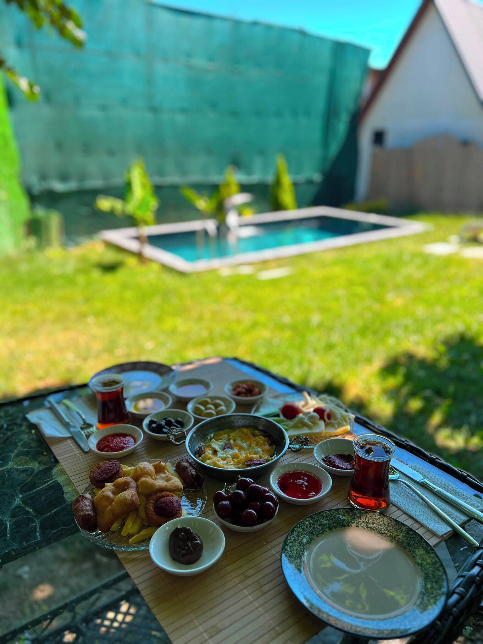 A picnic setup with a variety of foods and drinks on a table, a swimming pool in the background, and a grassy area.