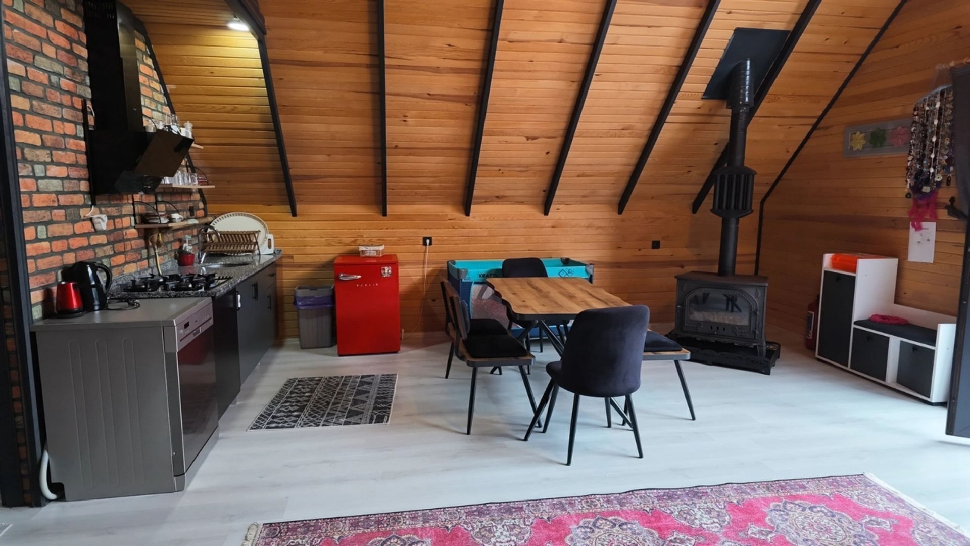 A cozy attic room with a kitchen, dining table, chairs, and a wood-burning stove.