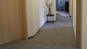 Un couloir recouvert de moquette, avec des portes en bois, une petite table sur laquelle est posé un vase, et, au fond, on aperçoit une pièce avec un bureau et une chaise.