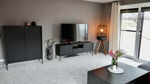 Un salon moderne avec un téléviseur à écran plat, un meuble de couleur sombre, une petite table ornée d’un vase de fleurs et une grande fenêtre avec des rideaux.