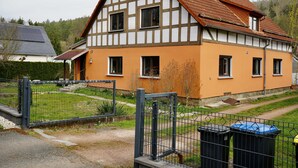 Une maison à deux étages, avec un toit en pente, une cheminée bien visible et une cour clôturée.