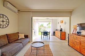 A living room with a sofa, a coffee table, and a dining area with chairs and a table. There is a kitchen area with cabinets and a TV.