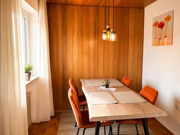 A dining area with a wooden table, orange chairs, a hanging light fixture, and a window with curtains.
