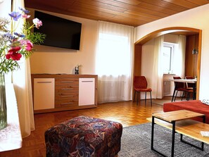 A living room with a wooden cabinet, a flat-screen TV, a patterned ottoman, a small table, and a red chair.