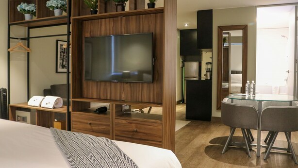 Ein modernes Hotelzimmer mit einem großen Bett, einem an einer Holzpaneele Wand montierten Fernseher, einem Glastisch mit Stühlen und einer kleinen Küchenzeile im Hintergrund.