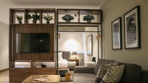 Ein modernes Wohnzimmer mit Sofa, Couchtisch und Fernseher. Pflanzen stehen auf Regalen, im Hintergrund ist ein Bett zu sehen.