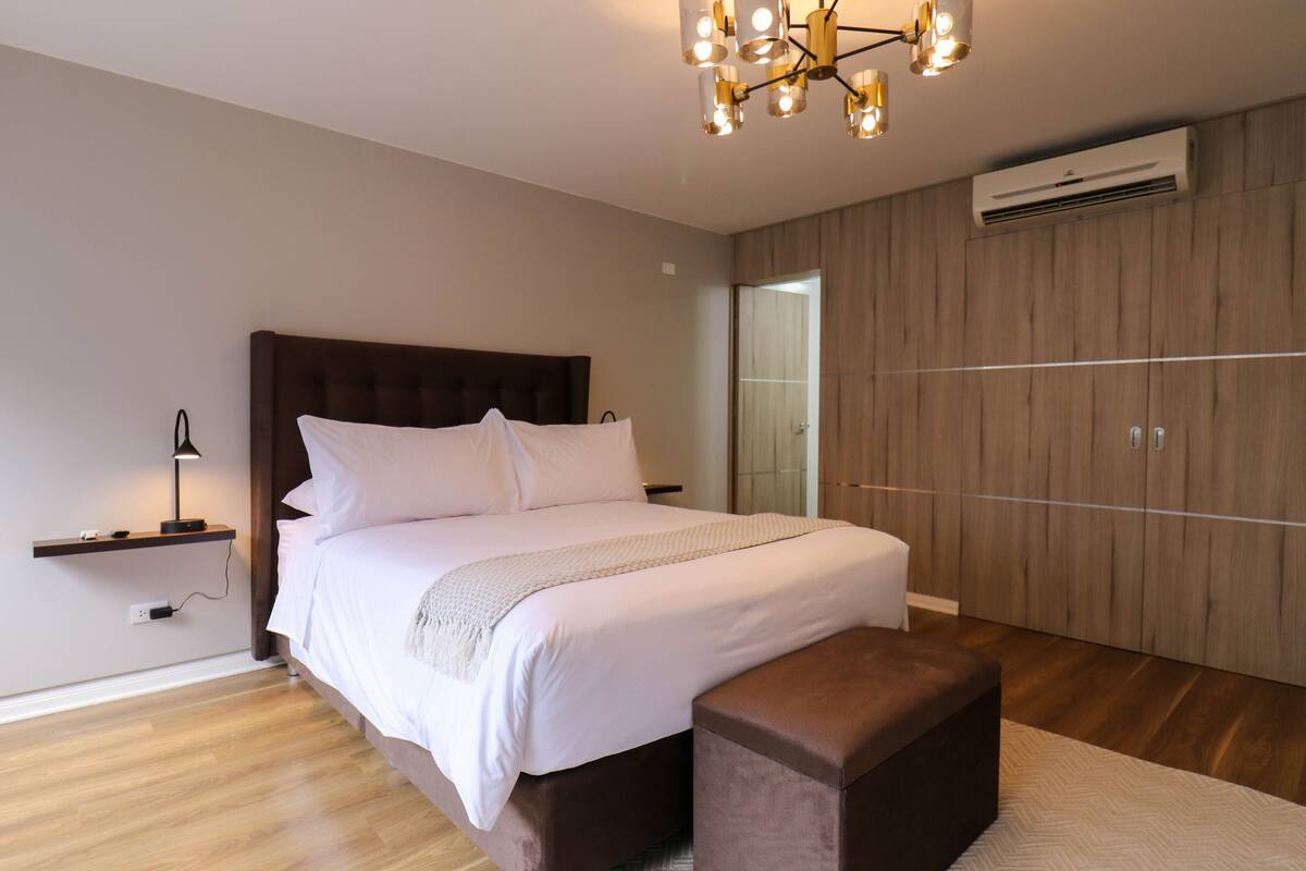 Quarto moderno com uma cama grande, uma mesa de cabeceira com abajur, guarda-roupa de madeira e unidade de ar condicionado na parede.