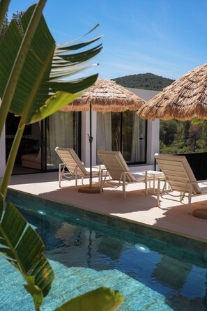 A poolside area with lounge chairs and thatched umbrellas, a clear pool, and a view of a forested hill. - Traumhafte Villa auf Ibiza Mitten im Grünen Gelegen, Strandnähe (Sant Josep de Sa Talaia)