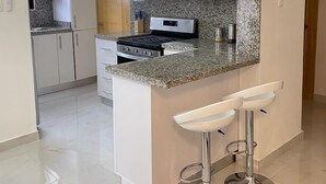 Cocina moderna con armarios blancos, encimera de granito, electrodomésticos de acero inoxidable y una isla central con dos taburetes de bar.