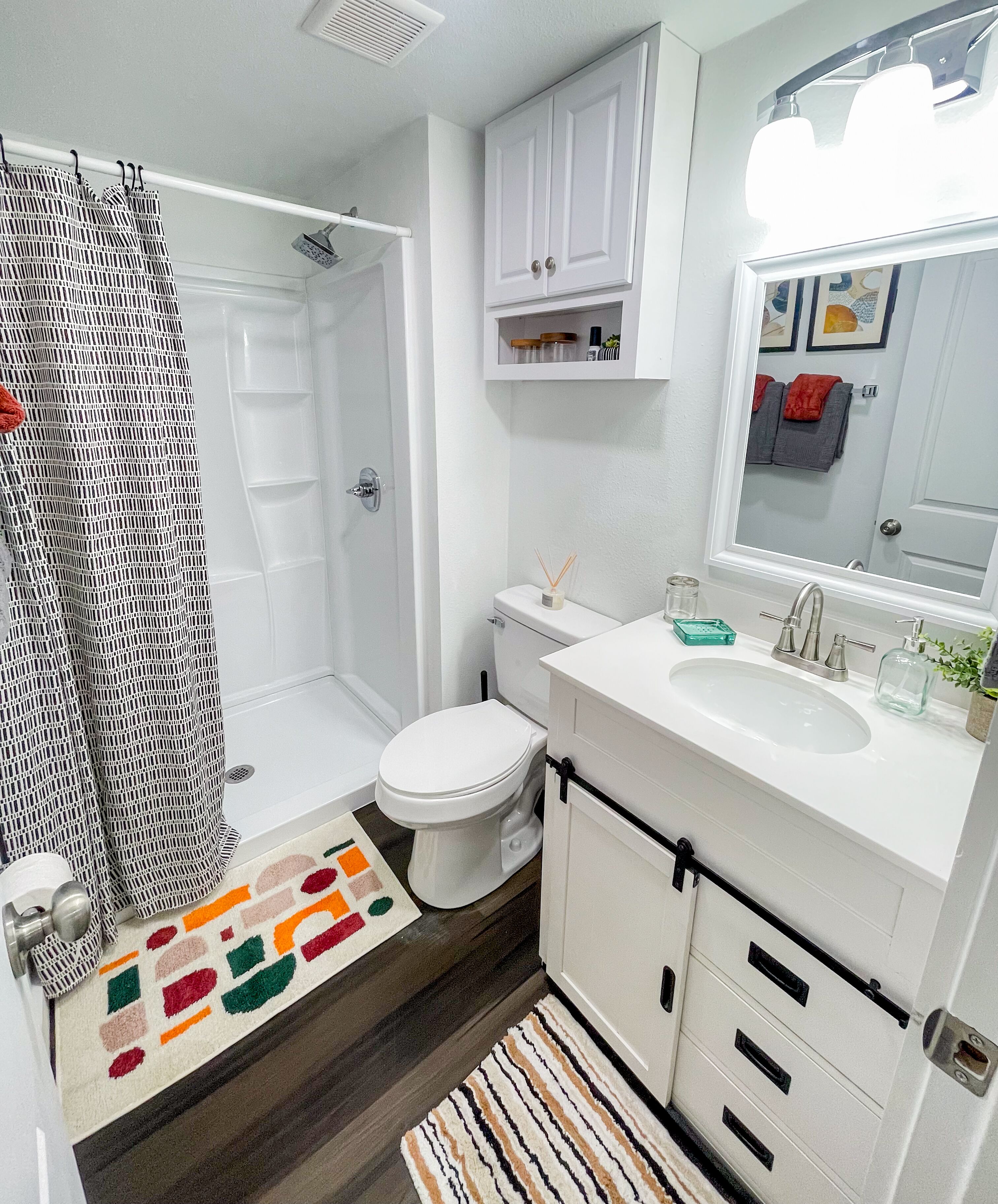 Une salle de bain avec des toilettes blanches, une douche avec un rideau à motifs, un lavabo avec un miroir au-dessus et un tapis coloré sur le sol.