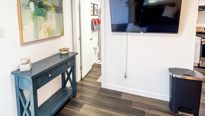 A modern living room with a dark wood floor, a blue console table, a flat-screen TV mounted on the wall, and a trash can. - Private Dallas/Bishop Arts Retreat  (Dallas)