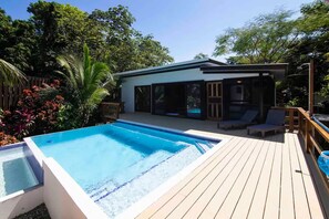 Un espace piscine extérieur moderne, doté d’une terrasse en bois, d’une chaise longue et d’un petit bâtiment avec de grandes fenêtres.