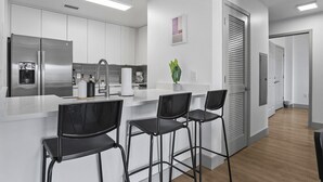 A modern kitchen with white cabinets, a stainless steel refrigerator, a sink, and a bar area with black chairs. - Beautiful Blue | Spacious, Free Parking, View, Gym (Charlotte)