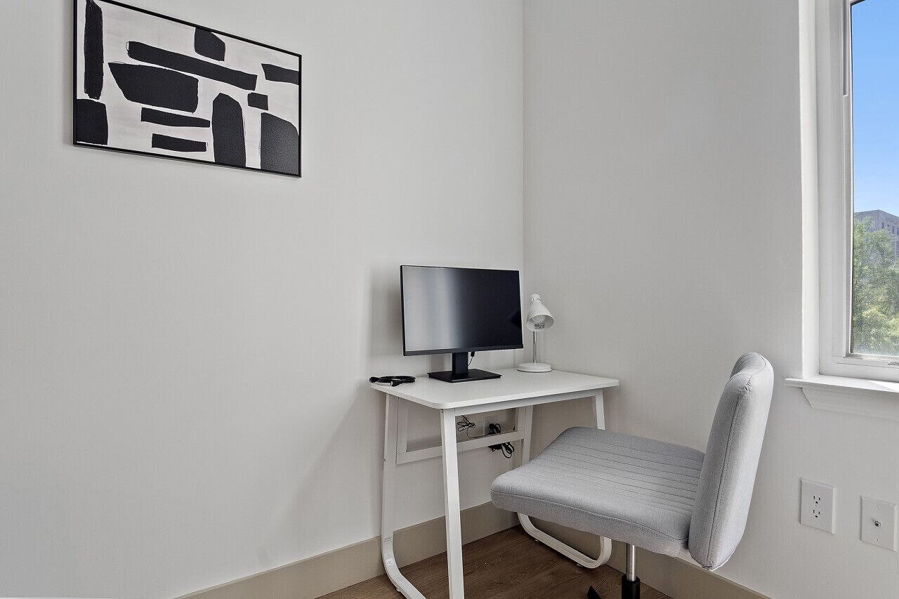 Una habitación pequeña y blanca con un escritorio, una silla, un monitor de computadora y una lámpara. Se ve un ventanal con vistas a la vegetación.