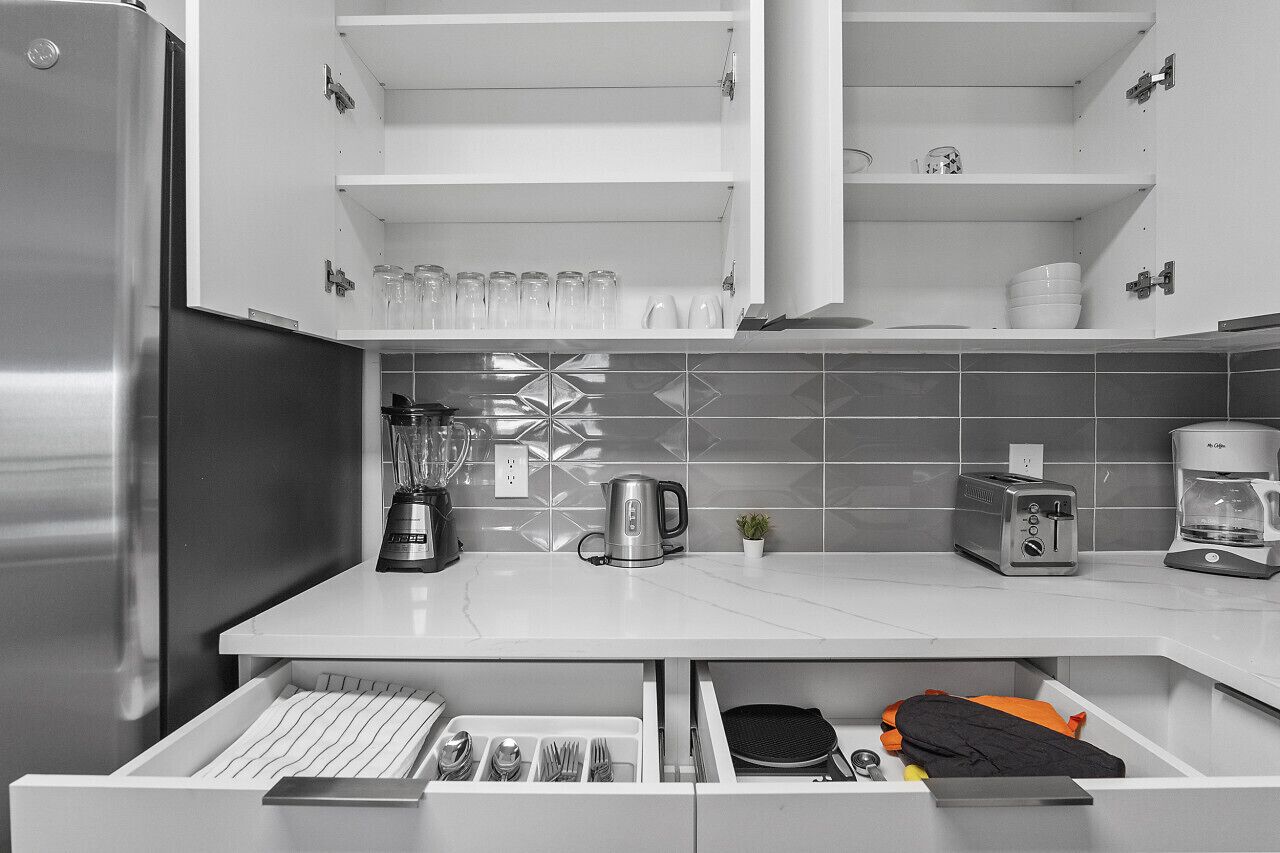 Una cocina con un refrigerador de acero inoxidable, una cafetera, un tostador y una tetera. Hay armarios sobre la encimera y un cajón debajo con utensilios.