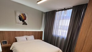 A modern hotel room with a wooden headboard, a large bed with white linens, a bedside table, a nightstand, a framed artwork on the wall, and a window with curtains. - Aerosuites (Bogotá)