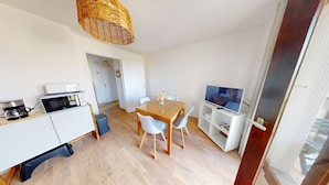 A compact living space with a dining table, chairs, a coffee machine, a TV, and a wicker hanging light fixture. - Appartement L'escalade Marine (Ouistreham)