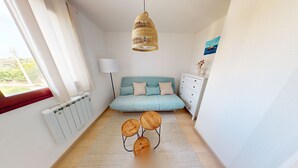 A modern bedroom with a light blue sofa, a hanging rattan light fixture, a white dresser, and a round wooden coffee table. - Appartement L'escalade Marine (Ouistreham)