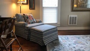 A cozy living room with a checkered sofa, a wooden chair, a floor lamp, a large window with blinds, a radiator, and a framed picture on the wall. - The Alexandria House #3
 (Jackson)