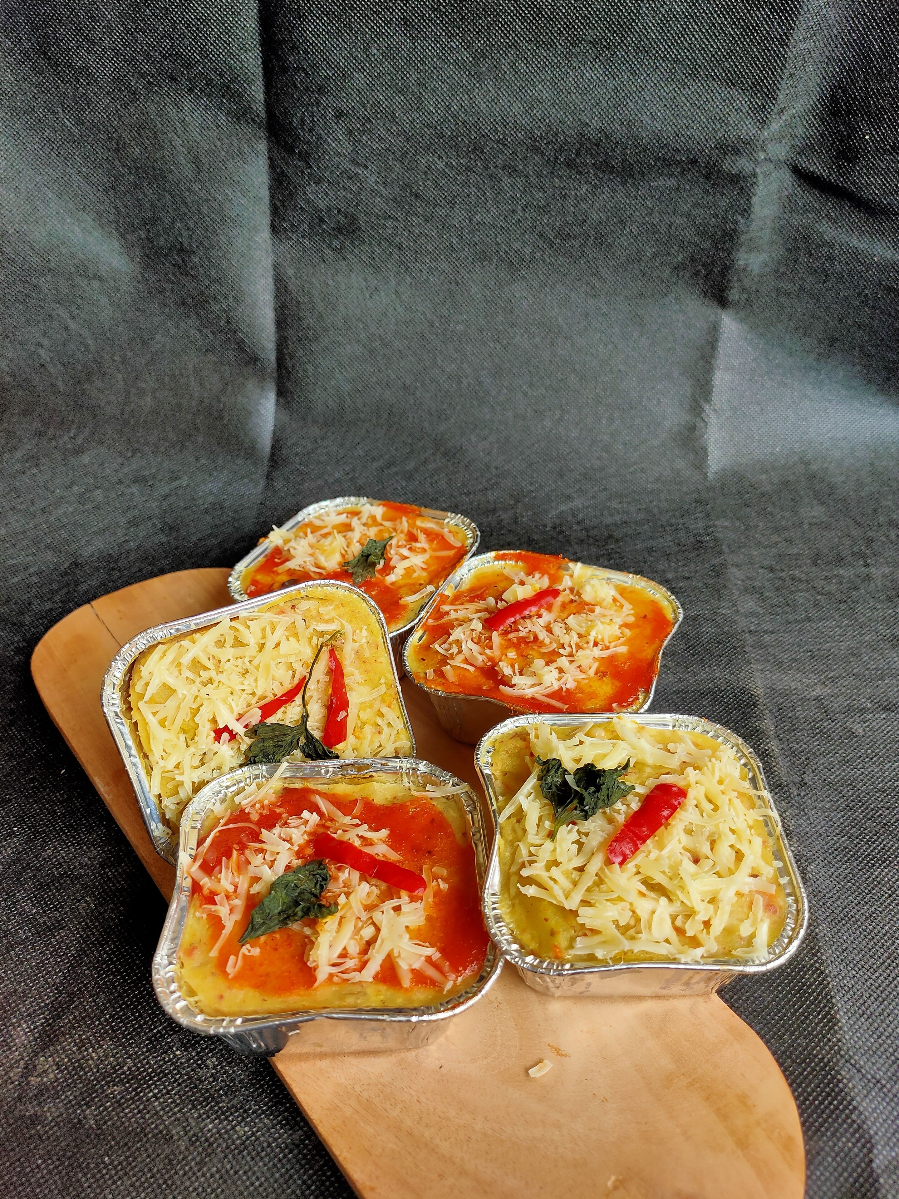 Five aluminum trays with food, placed on a wooden board.