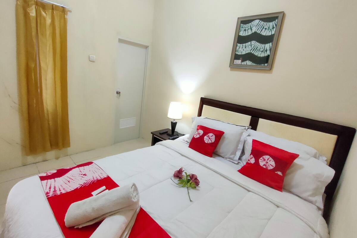 A neatly made bed with red and white bedding, a single red pillow, and a small bouquet of flowers.