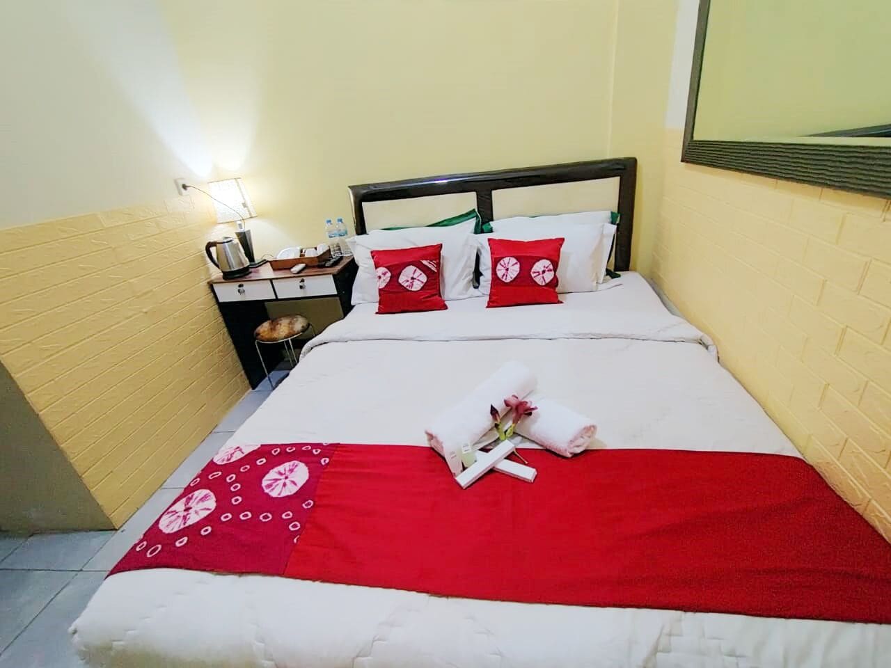 A neatly made bed with red and white bedding, a small bedside table with a kettle and bottles, and a wall-mounted mirror.