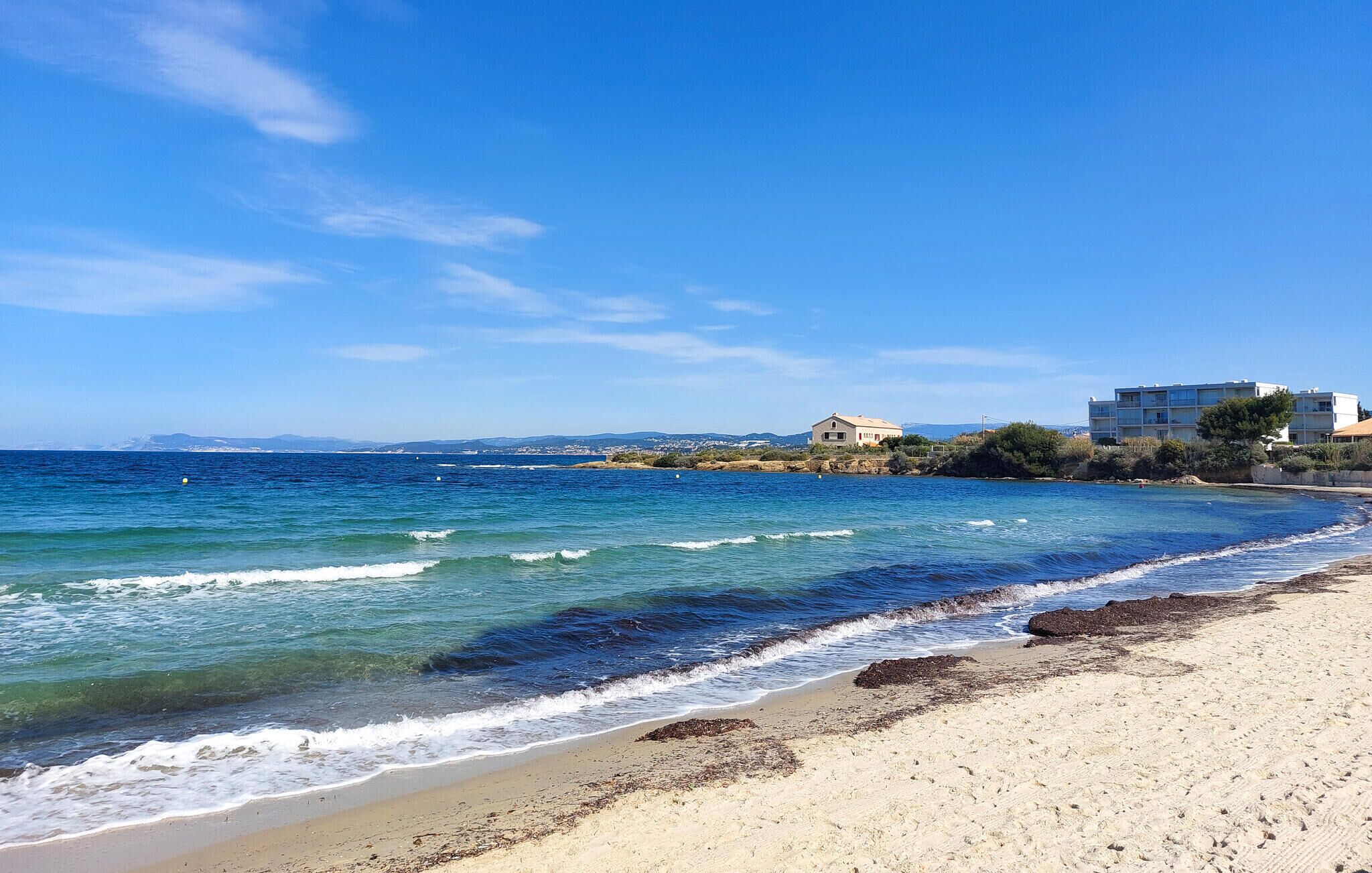 Une plage avec une eau d’un bleu limpide, un rivage sablonneux et des bâtiments en arrière-plan.