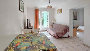 Una habitació amb taula de dinar, sofà, llit i cadira. Hi ha una finestra amb cortines, una làmpada i un quadre a la paret.