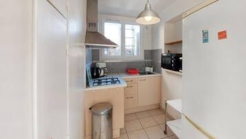 Een kleine keuken met een gasfornuis, een magnetron en een spoelbak. Boven de spoelbak is een raam en ernaast een vuilnisbak.