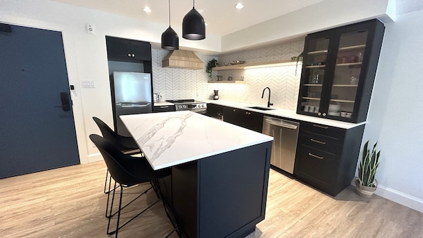 A modern kitchen with a central island, black bar stools, a built-in oven, and a dishwasher. - Le Héron - Studios Nature (Magog)