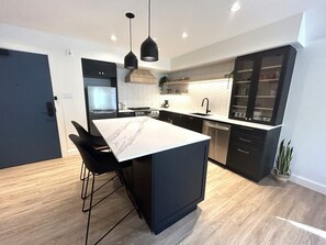 A modern kitchen with a central island, black bar stools, a built-in oven, and a dishwasher. - Le Héron - Studios Nature (Magog)