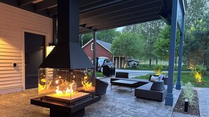 An outdoor patio with a modern fire pit, seating area, and a covered section with a barbecue grill. - Le Héron - Studios Nature (Magog)